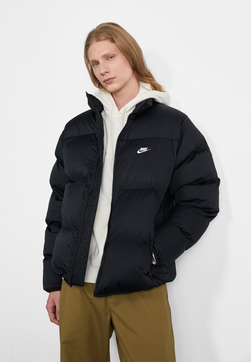 Sort pufferjakke med quiltet design, lynlås lukning og lille hvid Nike-logo på brystet. Båret over en lysefarvet hoodie og khakibukser.