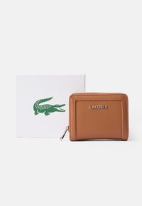 Brun skinnlommebok med teksturert overflate, med en sølvfarget glidelås og preget "LACOSTE" logo, ved siden av en hvit gaveeske med grønn krokodillelogo.