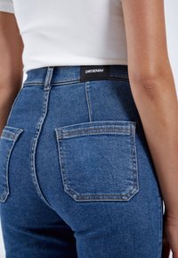 Jeans de mezclilla azul de talle alto con dos bolsillos traseros, textura desvanecida y una etiqueta negra de DRDENIM en la cinturilla.