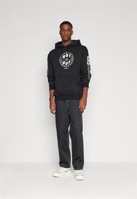 Svart hettegenser med grafisk print og kengurulomme, parret med rette mørke bukser og hvite sneakers. Enkelt design, casual passform.