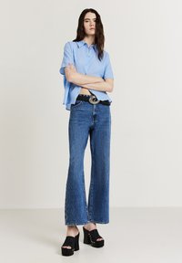 Donna che indossa una camicia azzurro chiaro a maniche corte con bottoni, jeans a vita alta a palazzo, una cintura intrecciata nera e sandali con tacco a piattaforma neri, in piedi con le braccia incrociate.