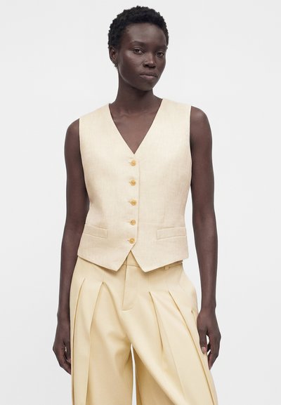 Σκοτεινόχρωμο άτομο που φοράει μπεζ αμάνικο γιλέκο με κουμπιά και μπεζ ψηλόμεσα πλισέ παντελόνια, στέκεται μπροστά σε απλό λευκό φόντο.