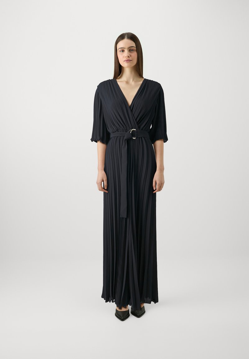 Abito maxi plissettato blu navy con scollatura a V avvolgente, maniche corte, vita con cintura e una consistenza liscia. Tacchi neri a punta completano il look.