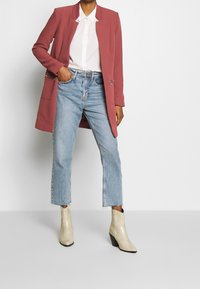 Růžový strukturovaný blazer přes bílou košili s knoflíky, doplněný světle modrými zkrácenými džíny a texturovanými hnědými kotníkovými botami.