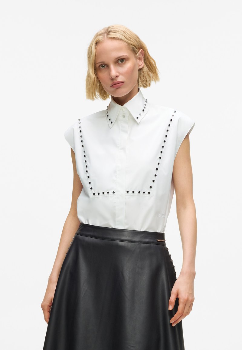 Blonde woman wearing sleeveless white shirt with black studs and black leather skirt, standing against white background.