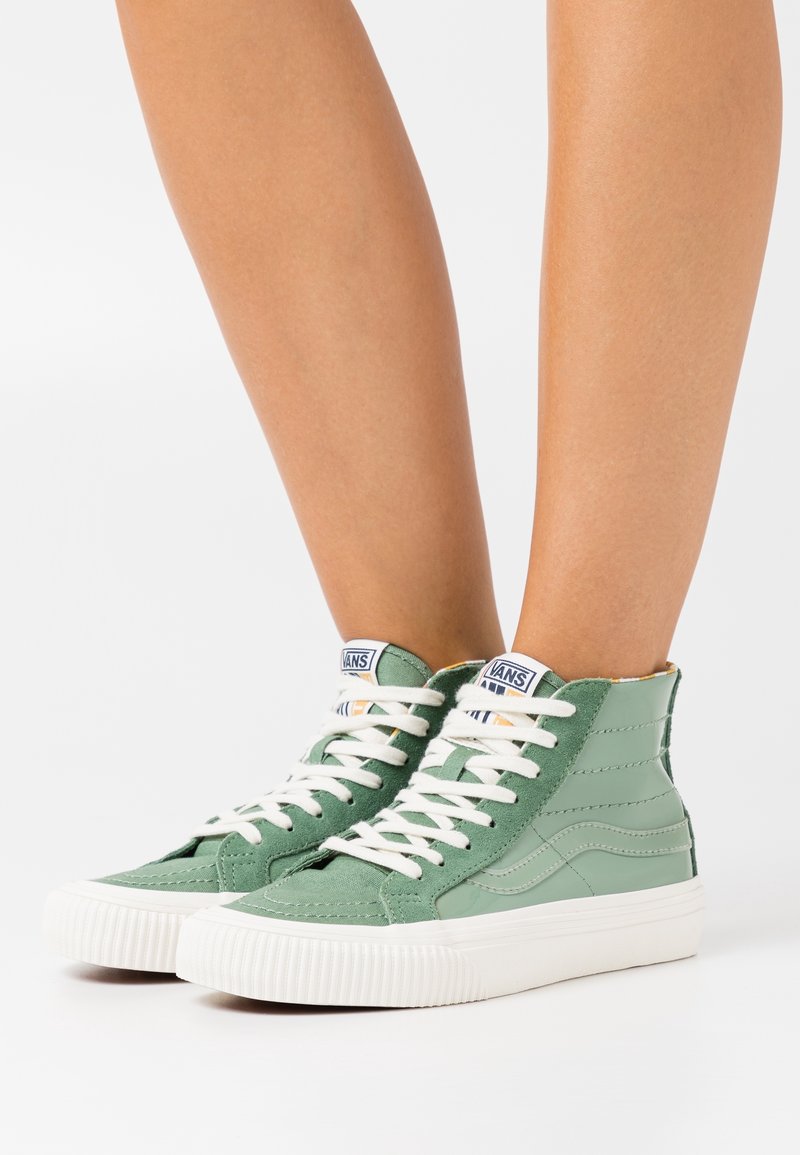 Ben med högt upp i Vans-sneakers i grön mocka och läder med vita snören och tjocka vita gummisulor på en vit bakgrund.