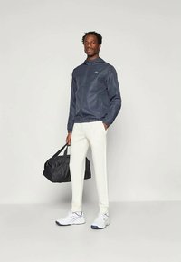 Casaco corta-vento azul-marinho com fecho na frente, bainha elástica; combinado com calças brancas e uma mala preta de lona. Modelo a usar ténis brancos.