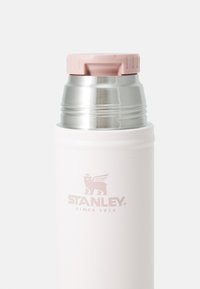 Thermos en acier inoxydable avec une finition blanche en poudre. Il présente un couvercle à vis rose et un logo imprimé en rose clair sur le corps.