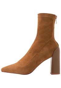 Bottine en daim marron avec un bout pointu, un talon bloc et une fermeture éclair à l'arrière, sur un fond blanc.