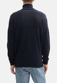 Granatowy sweter dzianinowy z wysokim kołnierzem, fakturowaną górną częścią oraz ściągaczami przy mankietach i u dołu. Posiada naszywkę na lewym rękawie.