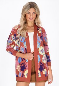Donna che indossa un cardigan floreale color ruggine sopra una maglietta bianca e una gonna marrone, con lunghi e ondulati capelli biondi, in piedi su uno sfondo bianco.