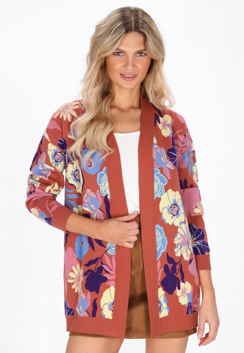 Donna che indossa un cardigan floreale color ruggine sopra una maglietta bianca e una gonna marrone, con lunghi e ondulati capelli biondi, in piedi su uno sfondo bianco.