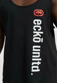 Mustat topit, jotka on valmistettu puuvillasta, jossa on pystysuora valkoinen teksti "eckō unlt." sivussa ja pieni punainen logo, jossa on sarvikuono design.