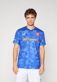 Modrý fotbalový dres s krátkým rukávem, který má pixelovaný vzor, výstřih do V a logo na hrudi, s bílým textem sponzora umístěným na prominentním místě.