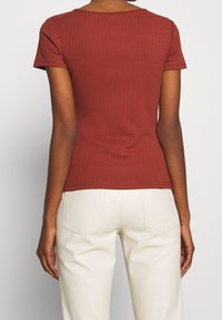 Žebrovaný, rezavě zbarvený top s krátkými rukávy a kulatým výstřihem, zobrazený ze zadní strany, spárovaný s krémově zbarvenými kalhotami.