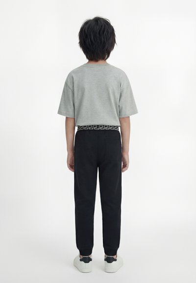 Παιδί που στέκεται με την πλάτη στραμμένη, φορώντας γκρι κοντομάνικο μπλουζάκι, μαύρο παντελόνι με σχέδιο στη μέση και λευκά αθλητικά παπούτσια με μαύρες λεπτομέρειες στη φτέρνα.