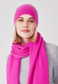 Cappello in maglia fucsia con texture a coste e sciarpa abbinata fucsia, entrambi realizzati in materiale morbido, indossati sopra un maglione grigio chiaro.