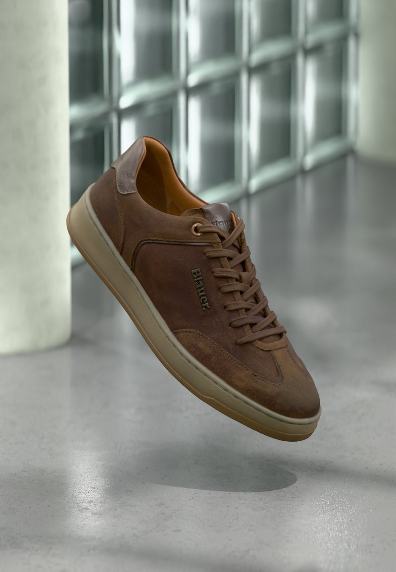 Zapatilla de piel marrón con puntera redondeada, superficie texturizada y cordones en tono similar. Presenta una suela de color claro y un logotipo sutil en el lateral.