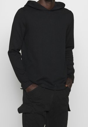 Hombre con sudadera negra lisa de manga larga y pantalones cargo negros con las manos en los bolsillos, sobre un fondo liso.
