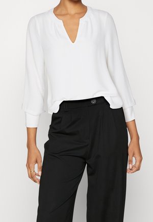 Kvinde iført en hvid bluse med lange ærmer og V-hals, stoppet ned i højtaljede sorte bukser, stående mod en ensfarvet baggrund.