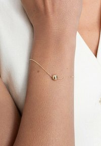 Złota bransoletka z cienkim łańcuchem i małym okrągłym zawieszką. Noszona na nadgarstku, prezentując gładką powierzchnię i delikatny wzór na jasnym tle.