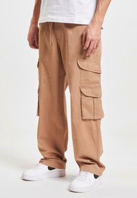 Beige cargobroek van katoen, voorzien van meerdere grote zijzakken, een losse pasvorm en een elastische taille. Gecombineerd met witte sneakers.