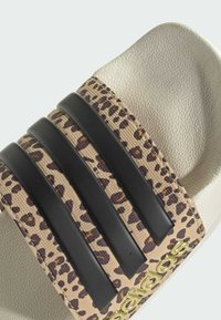 Snímek představuje béžový pásek s leopardím potiskem, černými gumovými pásky a zlatým logem. Podešev je texturovaná, v světle krémové barvě.