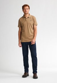 Polo color beige con cuello, mangas cortas y detalle de logo, combinado con jeans de mezclilla azul oscuro y zapatos marrones con cordones.