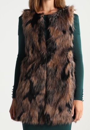 Kobieta ubrana w bezrękawnik z brązowo-czarnym wzorem z futra ekologicznego na ciemnozielonej sukience z długim rękawem, stojąca na jednolitym tle.