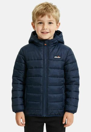 Ragazzo giovane con capelli biondi che indossa una giacca imbottita blu navy con cappuccio e zip frontale, sorridente verso la macchina fotografica.