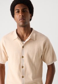 Kortærmet skjorte med knapper i lys beige, struktureret stof med subtile mønstre, der har brune knapper og et klassisk kravedesign.