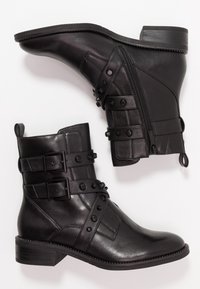 Botines de cuero negro con correas ajustables, tachuelas decorativas, cremallera lateral y un tacón bajo y apilado. Textura suave en toda su superficie.