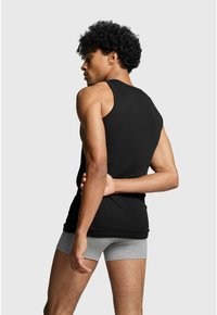 Black cotton tank top, sleeveless design, smooth texture, fitted shape. Styled with gray boxer briefs. Model shown from the back.