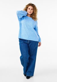 Hellblau gestrickter Pullover mit entspannter Passform und überschnittenen Schultern, kombiniert mit dunkelblauen weit geschnittenen Jeans und schwarzen Schuhen.