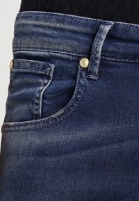 Primo piano di una tasca di jeans in denim blu scuro con cuciture visibili, passante per cintura e dettaglio in rivetto metallico.