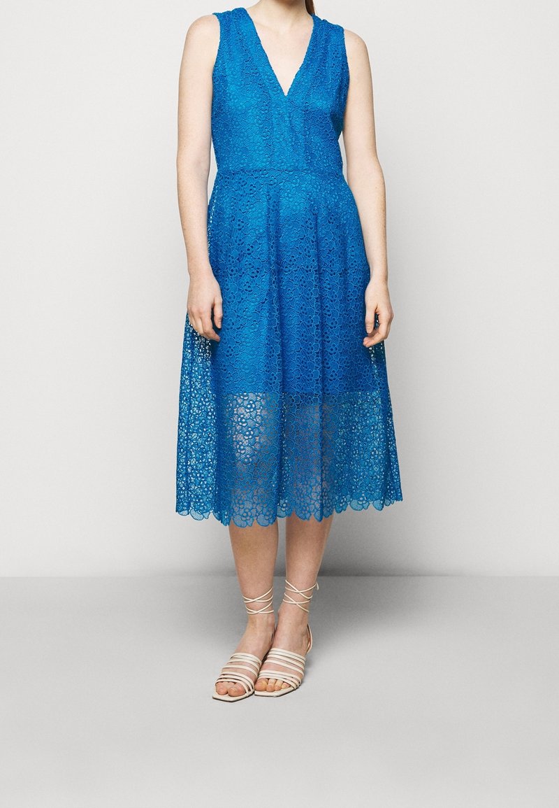 Robe en dentelle bleue avec décolleté en V, design sans manches et jupe jusqu'aux genoux. Présente un ourlet festonné et est assortie de sandales beiges à lanières.