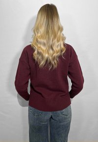 Maglione bordeaux a maglia con orlo e maniche a costine, abbinato a jeans azzurri a vita alta. Modella di spalle. Capelli biondi ondulati visibili.