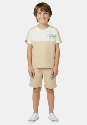 Garçon souriant portant un t-shirt et un short beige et blanc Lee Cooper avec des baskets blanches, debout devant un fond uni.