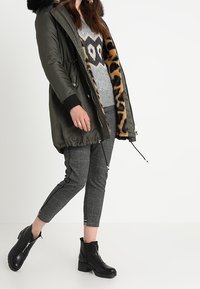Olivově zelená parka s umělým kožíškem na kapuci a leopardím potiskem uvnitř, spárovaná se šedým svetr s designem sovy a vzorovanými kalhotami. Černé kotníkové boty.