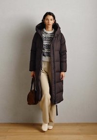 Manteau matelassé long marron avec capuche, porté sur un pull à motif. Le modèle porte un pantalon beige et des bottines blanches, tenant un sac marron.