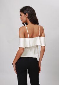 Haut blanc sans manches avec des accentuations volantes, texture plissée et bretelles réglables, associé à un pantalon noir ajusté.