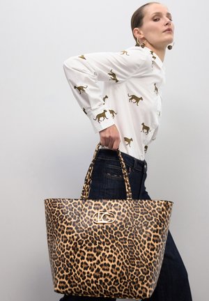 Mujer sosteniendo un bolso tote con estampado de leopardo, vistiendo una camisa blanca con estampados marrones de leopardo y pantalones oscuros contra un fondo liso.