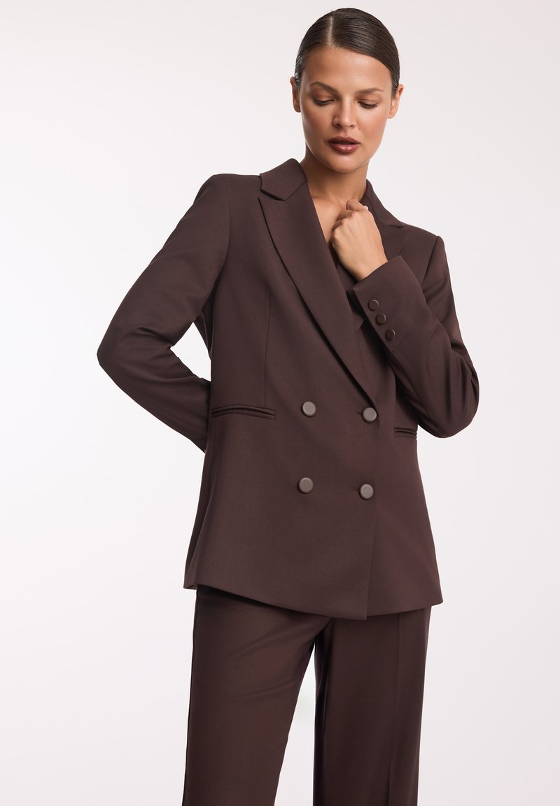 Brązowa marynarka dwurzędowa z wyciętym kołnierzem i subtelnymi detalami przy kieszeniach. Gładki materiał, dopasowany krój, zestawiona z pasującymi spodniami.