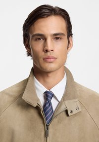 Μπεζ σατέν μπουφάν με όρθιο γιακά και ριμπ λεπτομέρειες, σε συνδυασμό με λευκό πουκάμισο και μπλε ριγωτή γραβάτα.