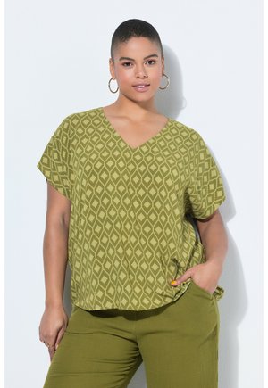 Kvinde med kort hår, iført guldfarvede øreringe i hoops, en grøn mønstret V-hals top og matchende grønne bukser, stående op ad en hvid væg.