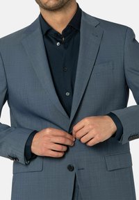 Hellgraue Sakko aus strukturiertem Stoff, mit Knopfverschluss und Reverskragen. Darunter ist ein schwarzes Hemd mit Knopfdetails sichtbar.