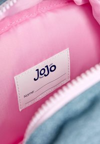 Szürke szövet táska belseje rózsaszín béléssel, egy fehér címkével, amelyen a "JoJo" felirat található, és van hely egy név kitöltésére.