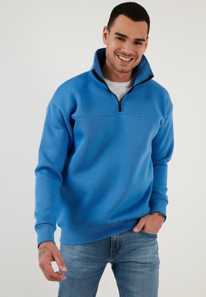 Lichtblauwe rits sweatshirt met een hoge kraag, ribbels bij de manchetten en onderkant. Gelaagde stof met een gladde afwerking. Gecombineerd met lichtblauwe jeans.