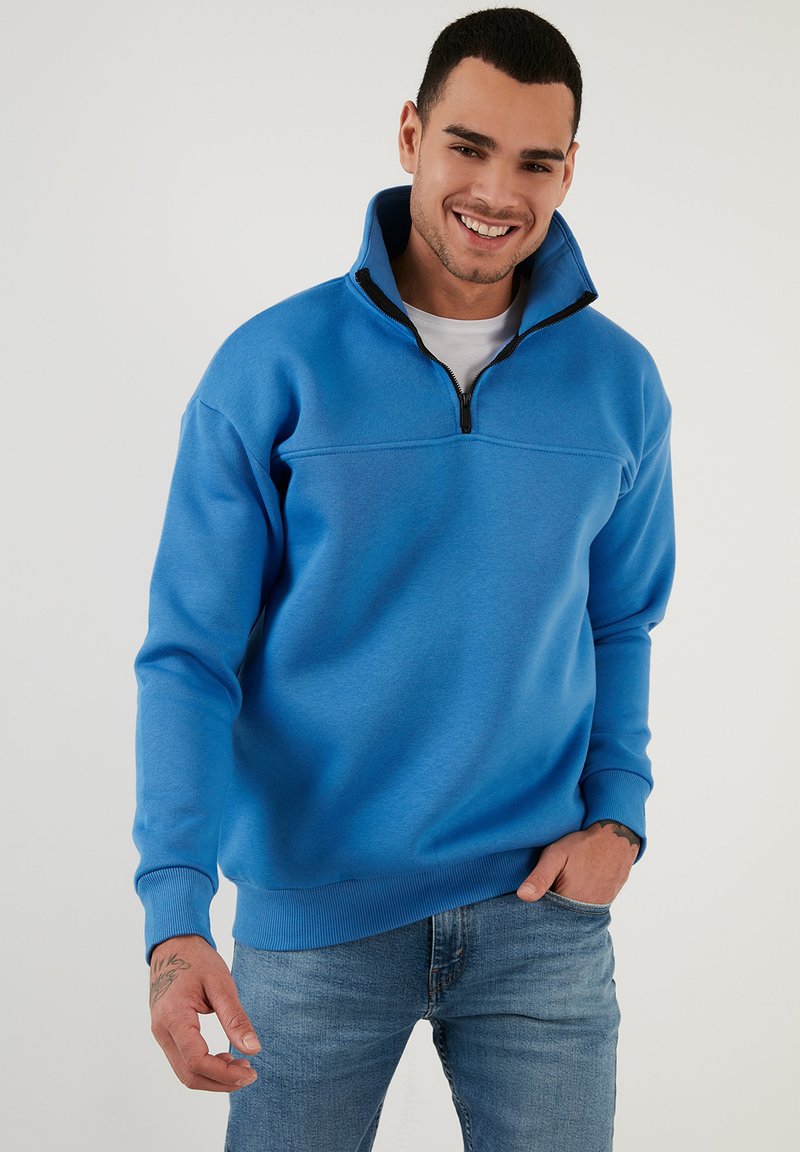 Felpa con zip azzurro chiaro, dotata di un colletto alto, maniche e orlo a coste. Tessuto testurizzato con un finish liscio. Abbinata a jeans azzurro chiaro.
