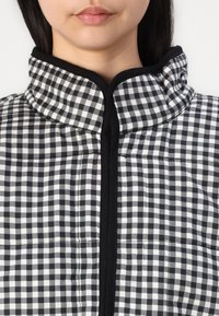 Jachetă matlasată cu model în carouri alb-negru, având un guler înalt, fermoar frontal și detalii negre de accent, oferind un aspect texturat.